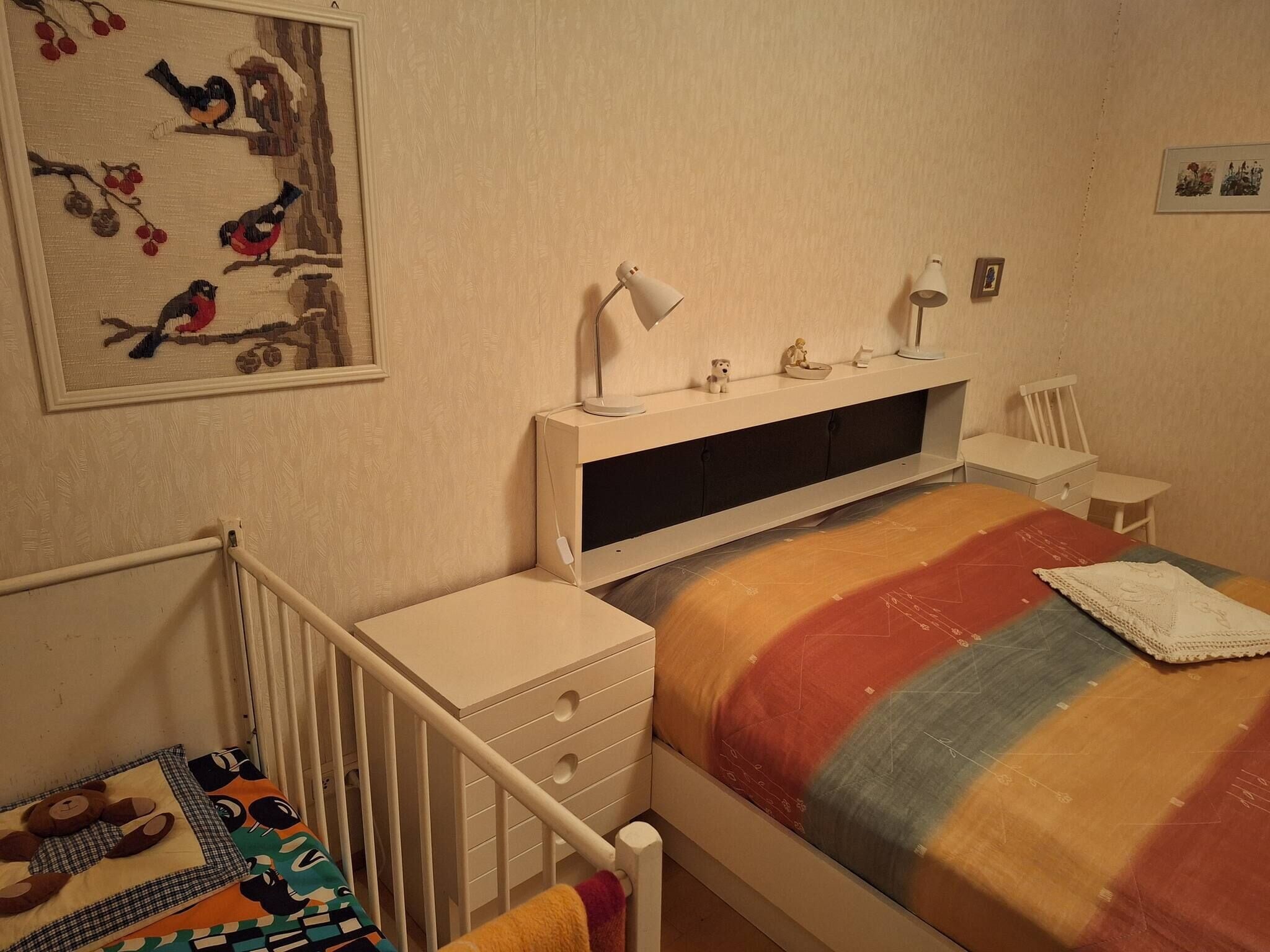 A bedroom with a bed, a nightstand, a desk, a chair, a lamp, and a wall-mounted shelf with decorative items.