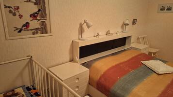 A bedroom with a bed, a nightstand, a desk, a chair, a lamp, and a wall-mounted shelf with decorative items.