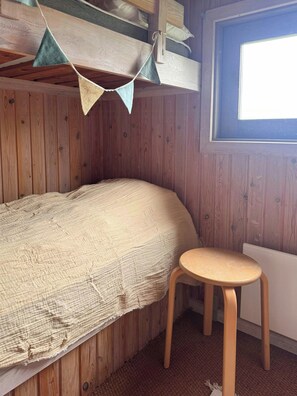 A single bed with a textured blanket, a small wooden stool, and a window with a view.
