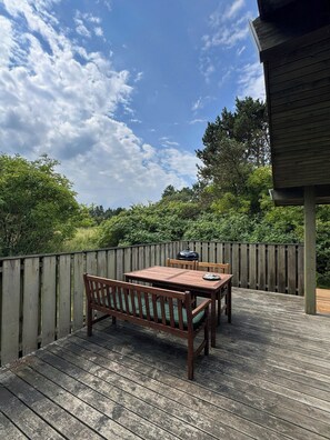 An outdoor wooden deck with a table and bench, surrounded by a wooden fence and dense greenery.