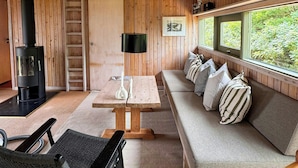 A cozy cabin interior with a wooden ceiling, a sofa with pillows, a table, a chair, a fireplace, and a window with a view of greenery. - House in Kettrup Bjerge near nature and the sea (Kettrup Bjerge)