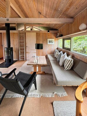 A cozy cabin interior with a wooden ceiling, a sofa with pillows, a table, a chair, a fireplace, and a window with a view of greenery.