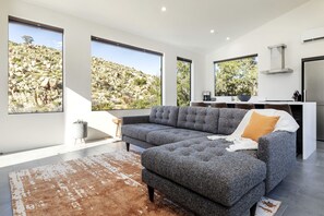 Ein modernes Wohnzimmer mit Ecksofa, großen Fenstern, einer Küchenzeile und Blick auf eine felsige Landschaft.