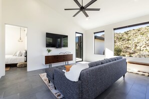 Uma sala de estar moderna com um sofá cinzento, uma televisão de ecrã plano, um aparador de madeira e uma janela grande com vista para um terreno rochoso.