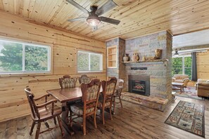 A wooden cabin interior with a dining table, chairs, a fireplace, and a ceiling fan.