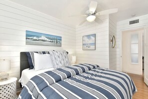 A bedroom with a bed, two wall-mounted paintings, a ceiling fan, and a door.