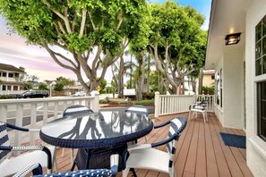 A covered outdoor patio with a round table and chairs, surrounded by white railings and large trees.