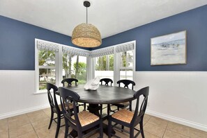 A dining area with a round table, six chairs, a woven pendant light, and a view through a bay window.