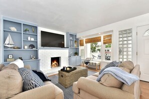 A living room with a white brick fireplace, blue shelving, a flat-screen TV, and a view of the outdoors.