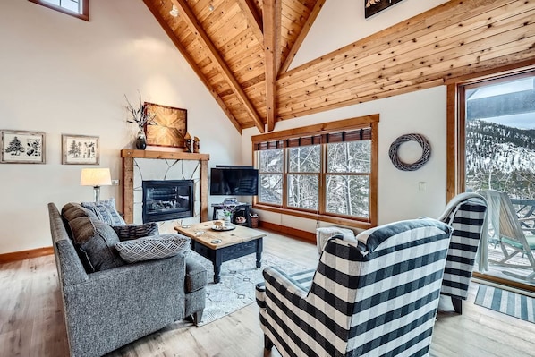 A cozy living room with a fireplace, a sofa, a striped armchair, and a large window with a view of snow-covered trees.