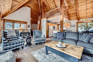 A cozy cabin interior with a living room, dining area, and kitchen, featuring wooden beams and a loft.