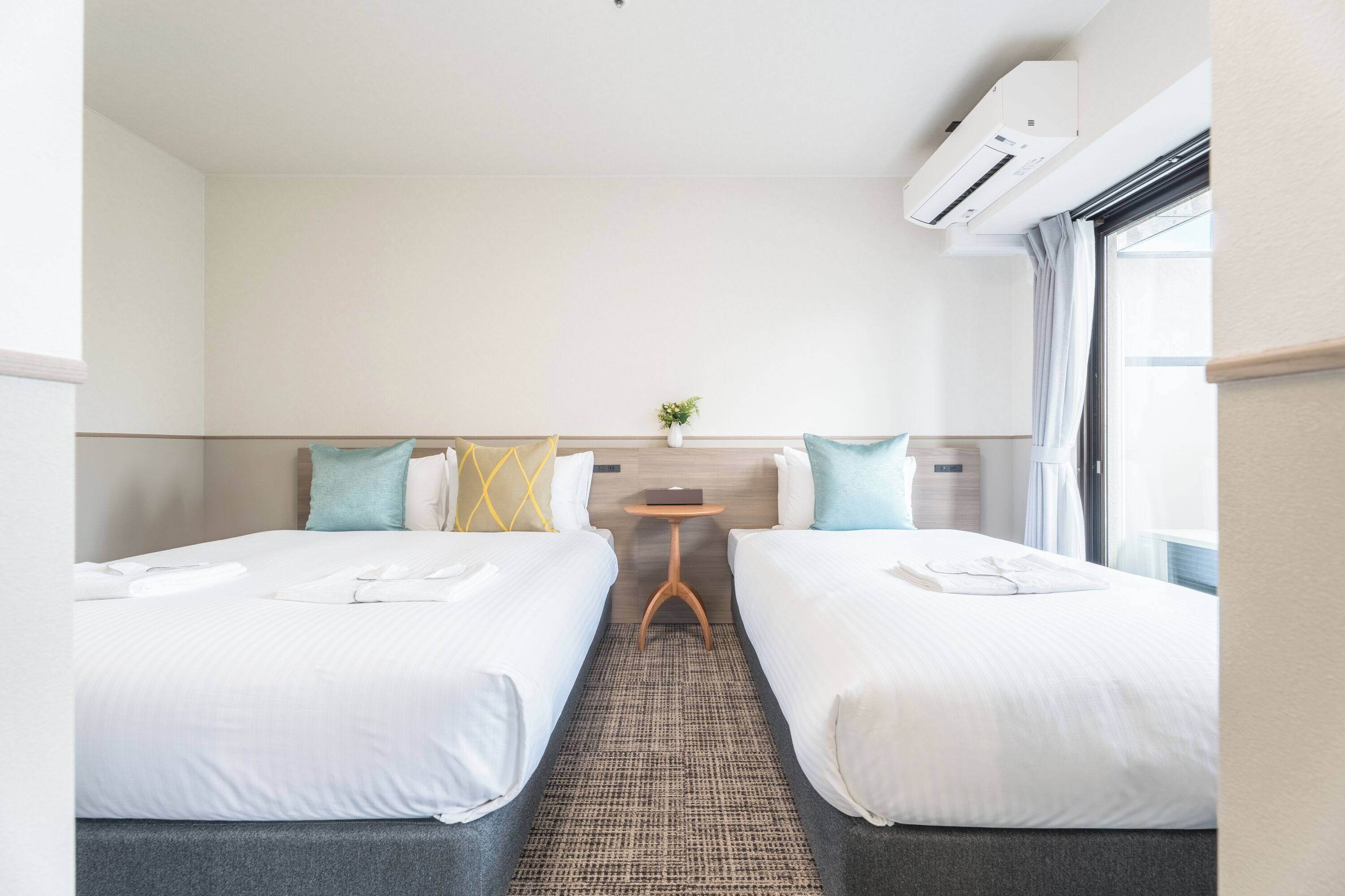 A hotel room with two beds, a small table, a plant, and an air conditioning unit.