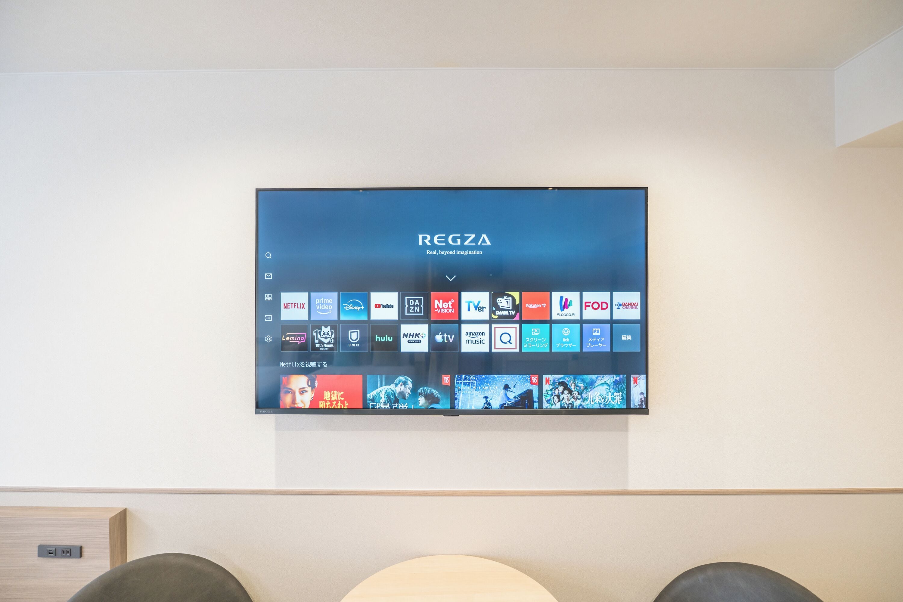 Ein an der Wand befestigter Fernseher zeigt verschiedene Streaming-Dienste und das Logo von 'REGZA' mit dem Slogan 'Real. Jenseits der Vorstellungskraft.'