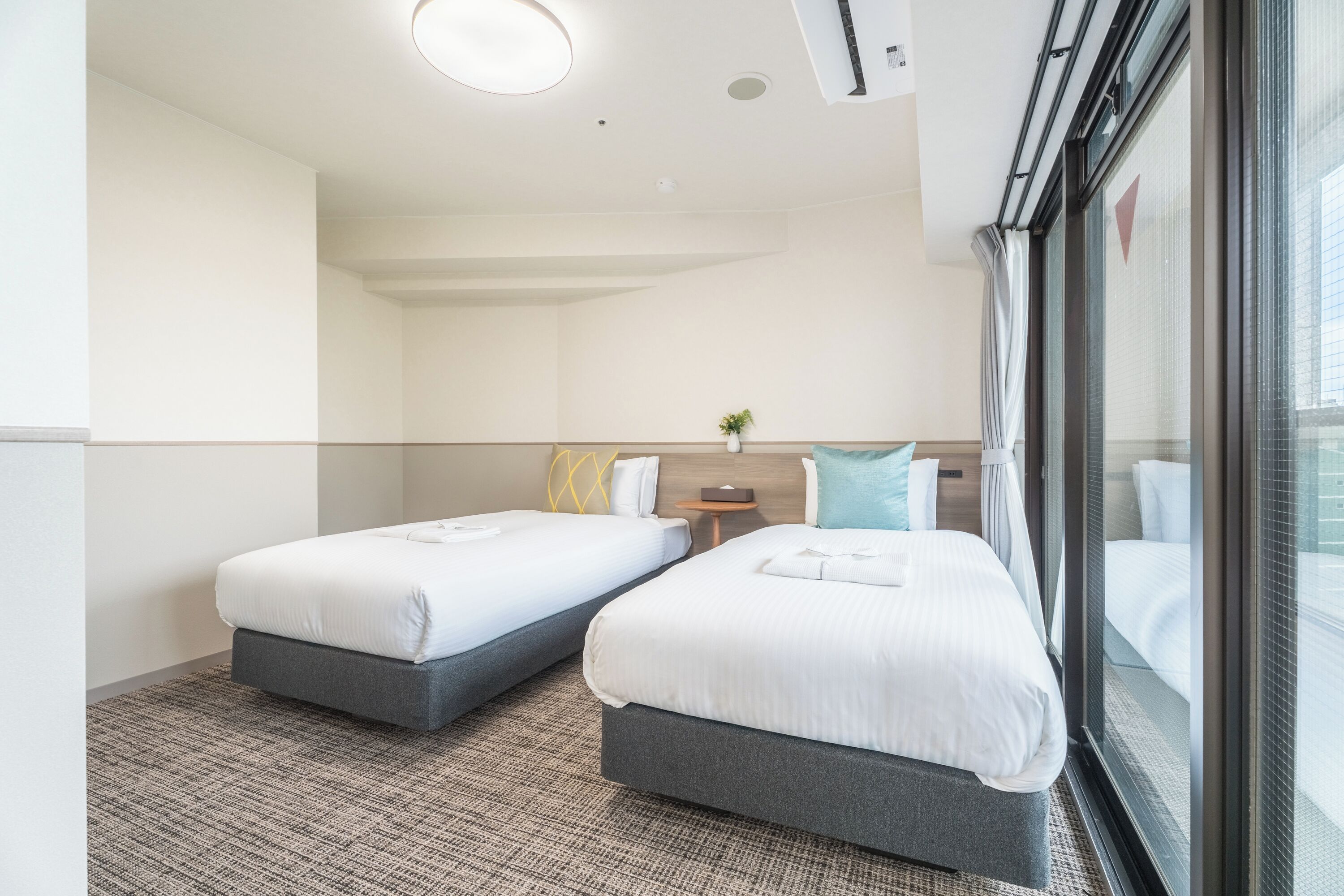 A hotel room with two beds, a small bedside table, a wall-mounted air conditioner, and a window with a view.