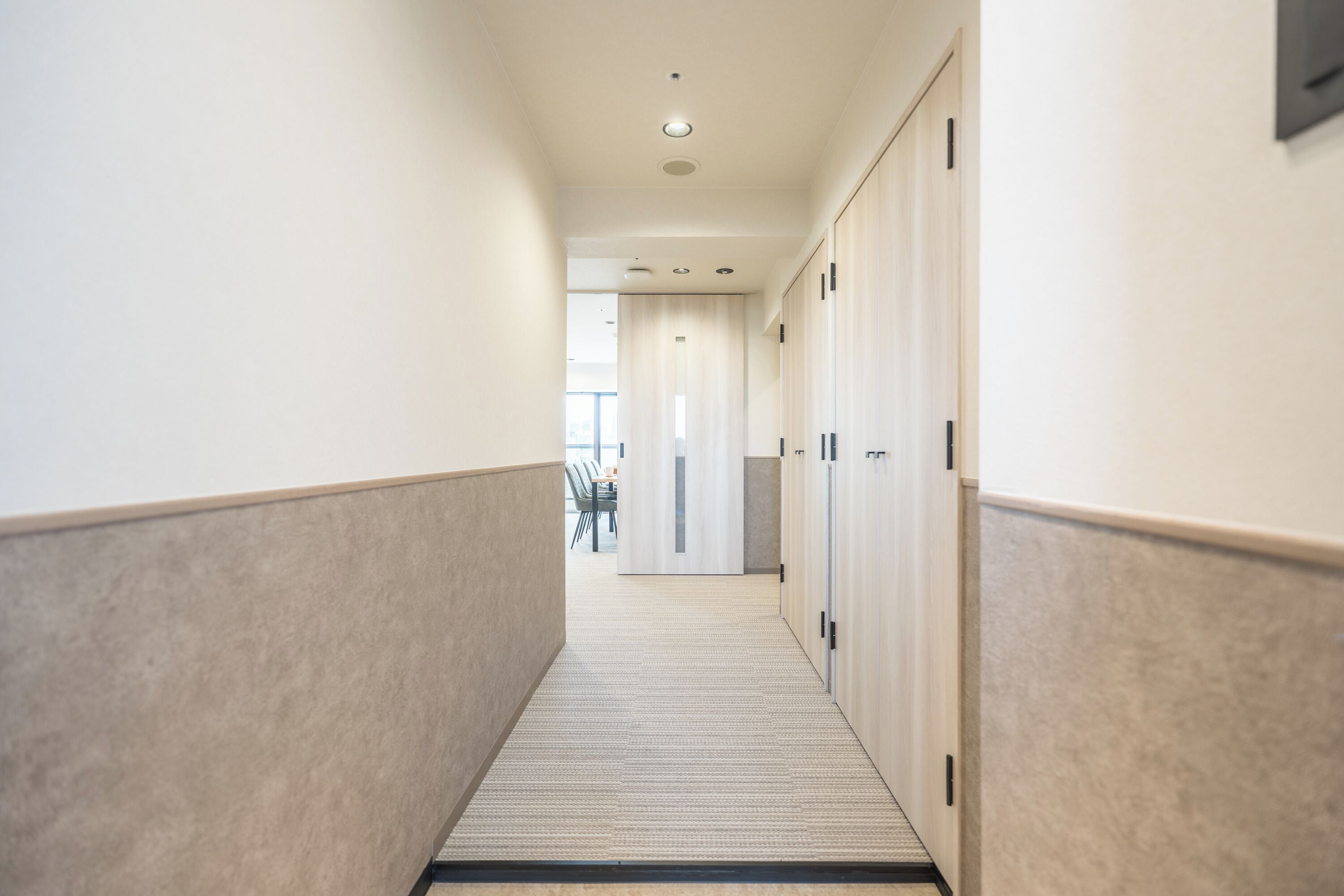 A hallway with lockers, carpeted floor, and a door at the end leading to a room.