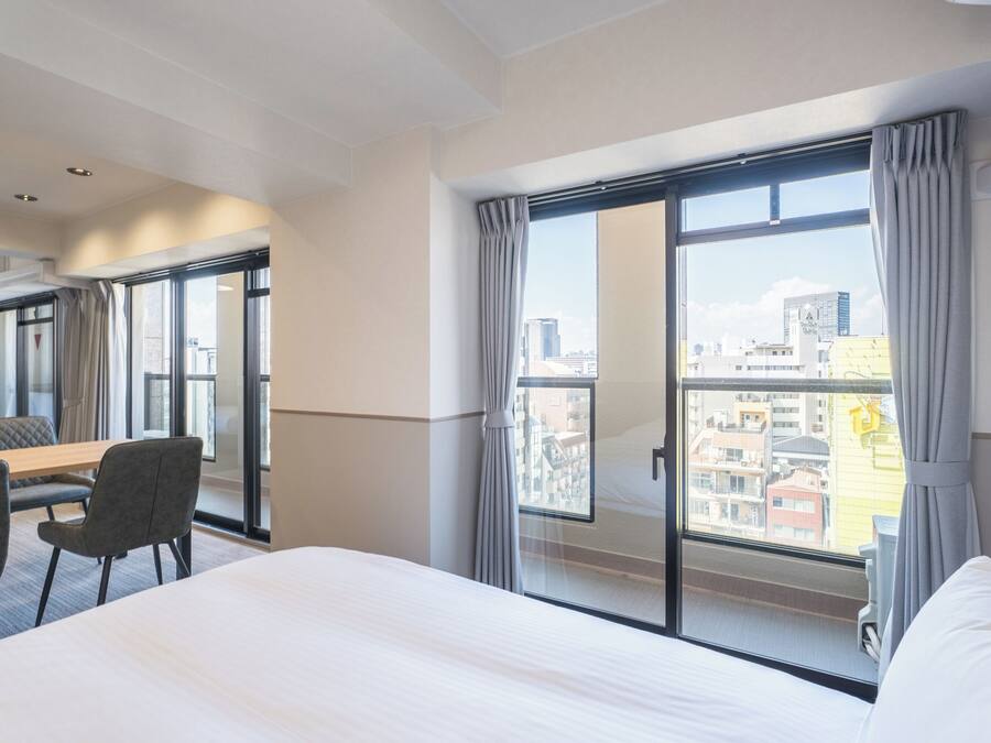 A hotel room with a bed, a dining table with chairs, and a large window with curtains.