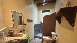 Um banheiro com pia verde, uma bacia de pedra, um espelho com moldura decorativa, vaso sanitário e área de chuveiro com parede de pedra.