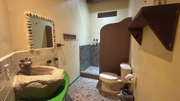 A bathroom with a green sink, a stone basin, a mirror with a decorative frame, a toilet, and a shower area with a stone wall.
