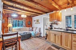Interior d'una cabana acollidora, amb sostre de fusta, un sofà, taula de dinar, zona de cuina i una finestra gran.