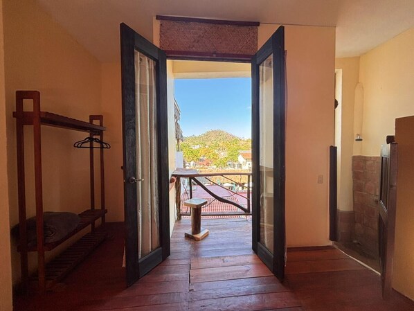 Uma porta aberta dá acesso a uma varanda com vista para uma colina e edifícios.