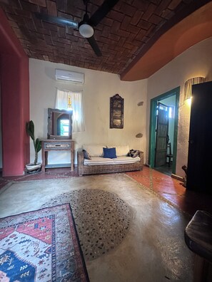 Una habitación con techo de ladrillo, un sofá con cojines, un tapete estampado, una planta en maceta y un cuadro colgado en la pared.