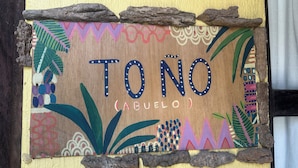 Uma placa de madeira com a inscrição 'TONO (ABUELO)' cercada por padrões coloridos e elementos naturais.