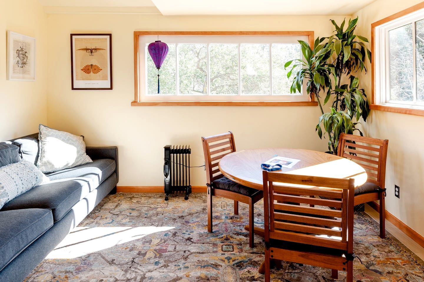 Ein gemütliches Wohnzimmer mit einer Couch, einem runden Holztisch mit Stühlen, einer Heizung und einem großen Fenster mit Blick auf Bäume.
