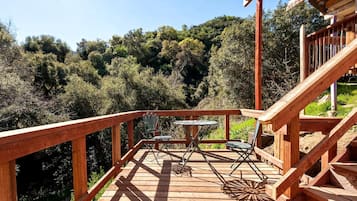 A wooden deck with a table and chairs, surrounded by a railing and overlooking a forested area.