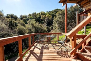 A wooden deck with a table and chairs, surrounded by a railing and overlooking a forested area.