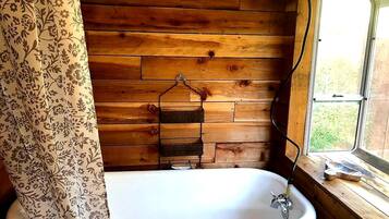 A wooden cabin bathroom with a white bathtub, a hanging towel rack, and a window with a view of greenery.