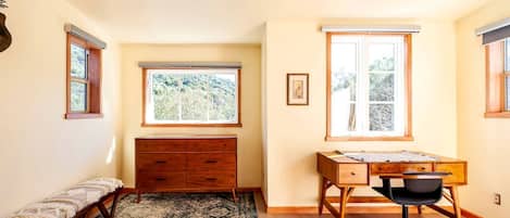 Un dormitorio con cama, escritorio, silla, cómoda y dos ventanas con vistas a zonas verdes.
