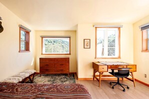 Ein Schlafzimmer mit einem Bett, einem Schreibtisch, einem Sessel, einer Kommode und zwei Fenstern mit Blick auf Grünflächen.