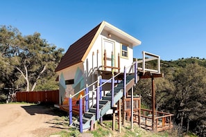 A small, single-story building with a sloped roof, a staircase leading to a balcony, and a fenced yard.