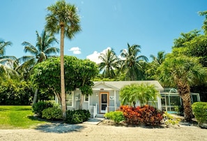 A single-story house with a front porch, surrounded by palm trees and tropical vegetation.