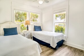 A bedroom with two beds, a nightstand with a lamp, and large windows with blinds.