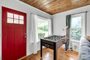 Une pièce avec une porte rouge, un baby-foot, deux fenêtres avec des rideaux et un plafond en bois.