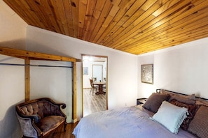 A bedroom with a wooden ceiling, a bed with multiple pillows, a chair, and a door leading to a dining area.