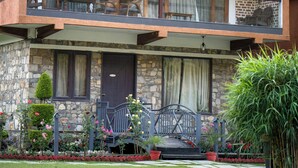Un edificio de dos pisos de piedra, con un balcón, entrada de piedra y un jardín con plantas en macetas.