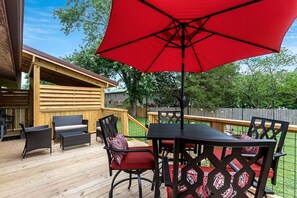 A wooden deck with outdoor furniture, a red umbrella, and a wooden fence in the background. - Cozy Cottage with Fire Pit and Barbecue in Pigeon Forge, Tennessee (Pigeon Forge)