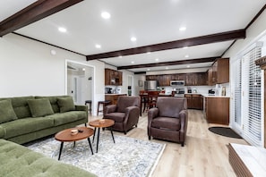 A spacious living room with a green sofa, two armchairs, and a coffee table. A kitchen with wooden cabinets and a dining area is visible in the background. - Cozy Cottage with Fire Pit and Barbecue in Pigeon Forge, Tennessee (Pigeon Forge)