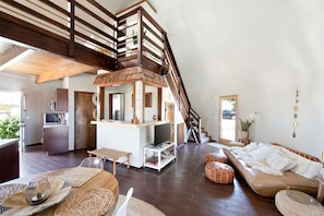 Un loft espaiós amb un sostre alt, una escala de fusta, una zona de cuina, una zona d'estar amb un sofà i un otomà, i una taula de menjador amb places de seient.