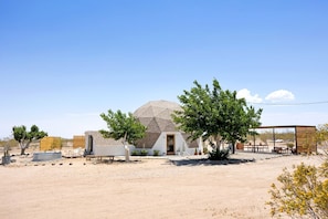 A desert landscape with a single building featuring a dome-like structure, surrounded by sparse vegetation and a clear sky.