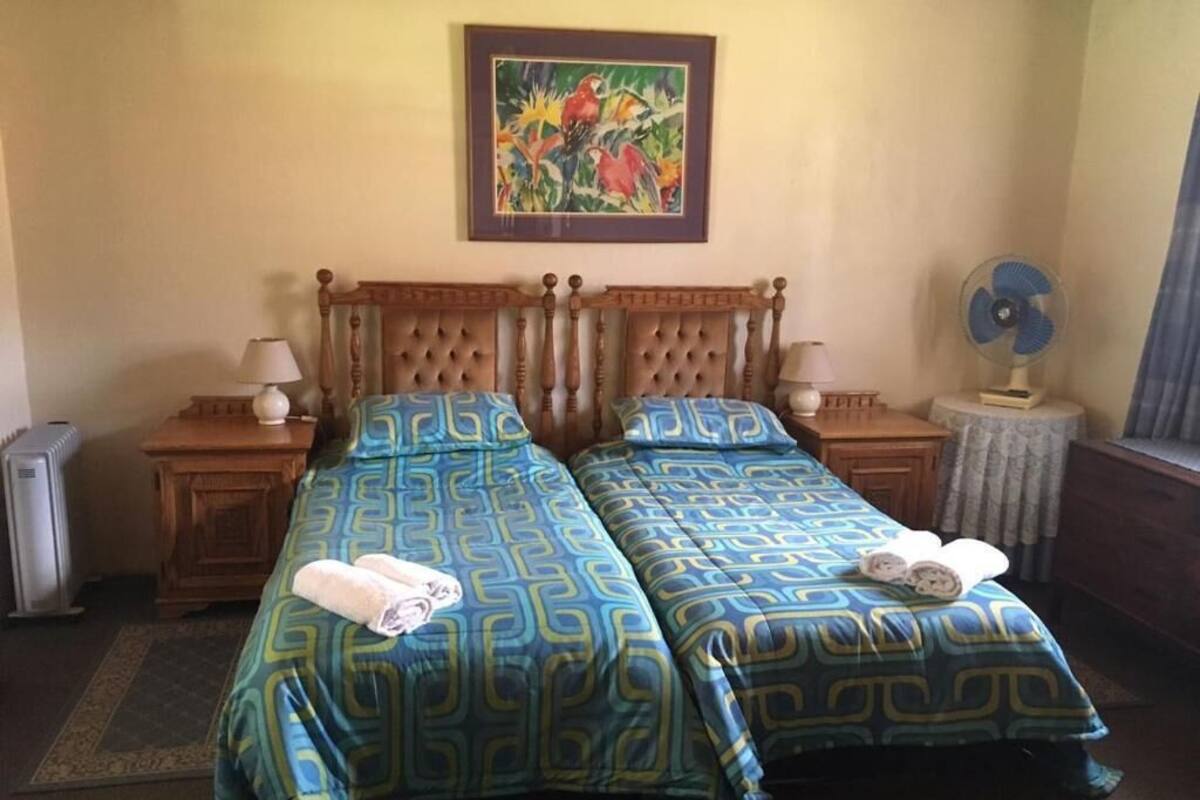 Ein Doppelbett mit passenden Kopfteilen, zwei Nachttische mit Lampen, ein gerahmtes Bild an der Wand und ein Standventilator.