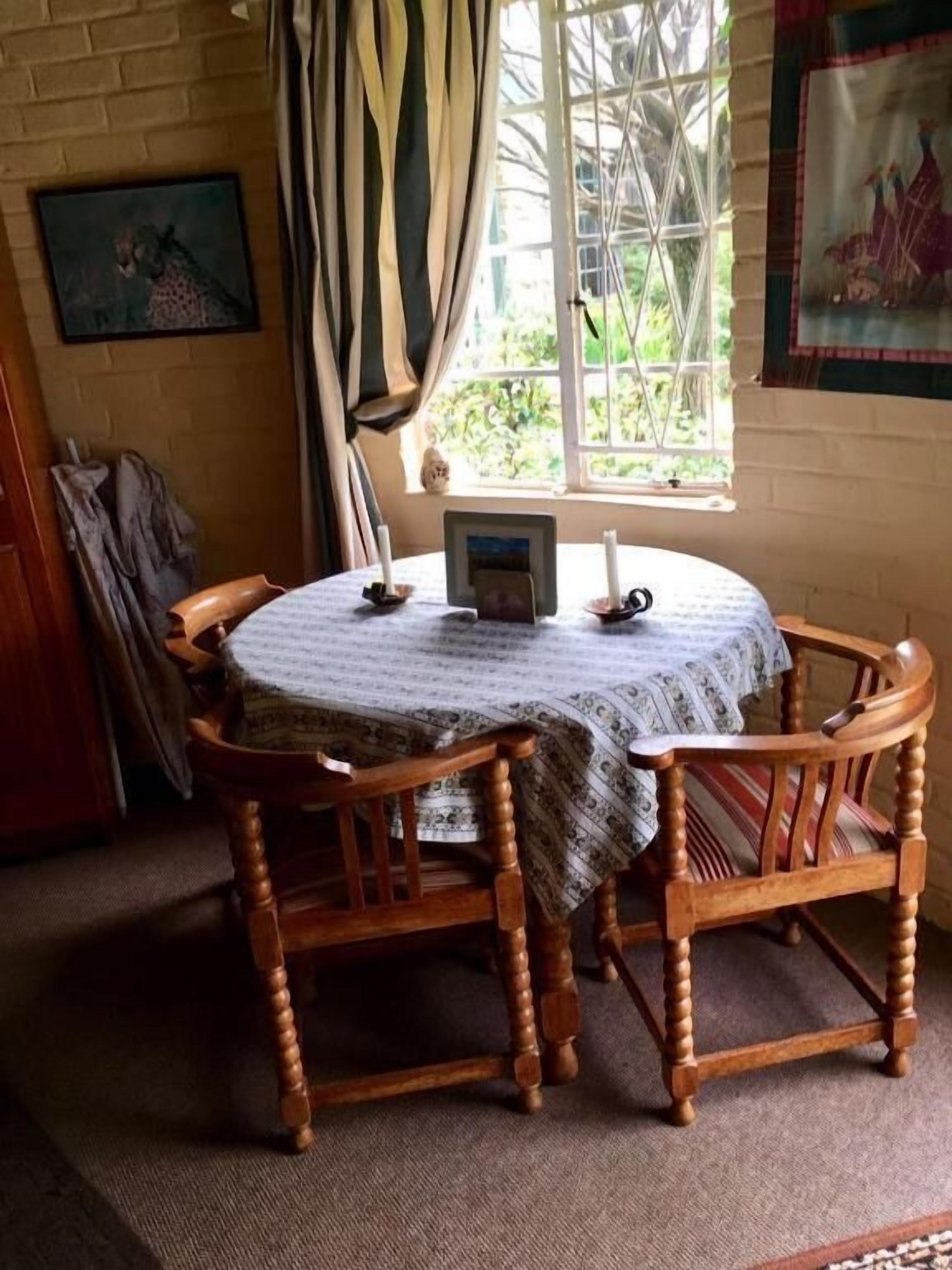 A round wooden table with a patterned tablecloth, surrounded by four wooden chairs, a window with curtains, and a framed picture on the wall.