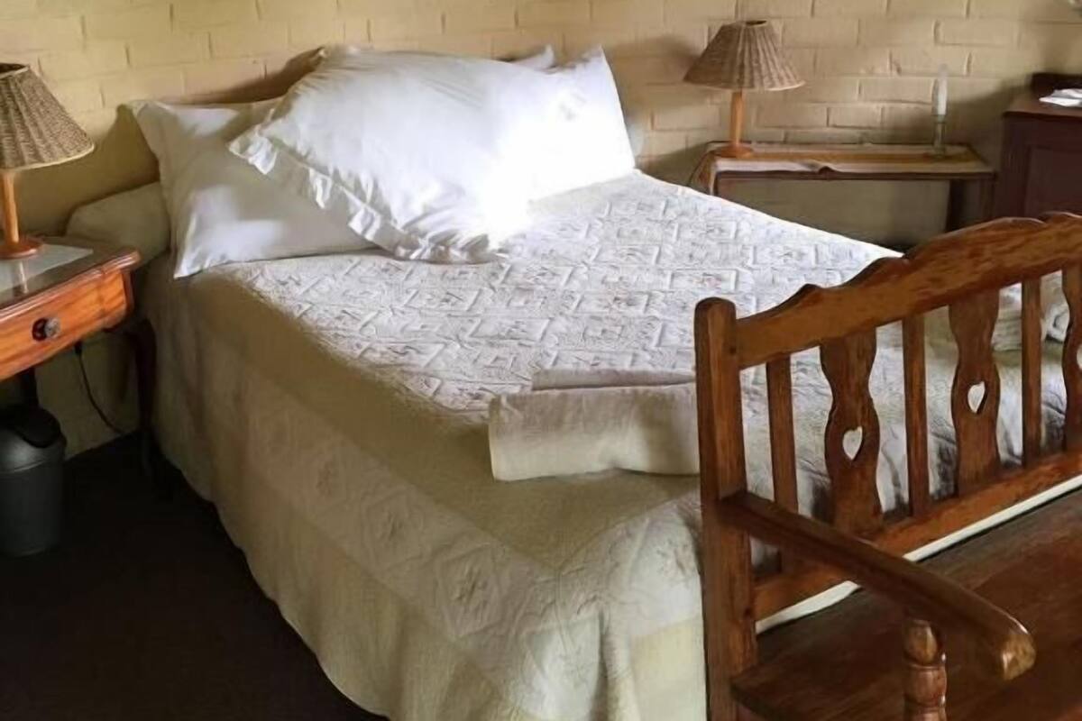 Ein Schlafzimmer mit Bett, Holz-Kopfteil, Nachttisch mit Lampe, einem an der Wand hängenden Bild und Teppichboden.