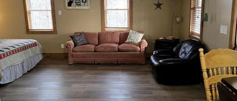 A living room with a ceiling fan, two windows, a sofa, a black leather chair, and a wooden chair.