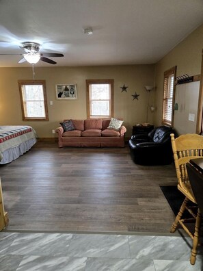 A living room with a ceiling fan, two windows, a sofa, a black leather chair, and a wooden chair.