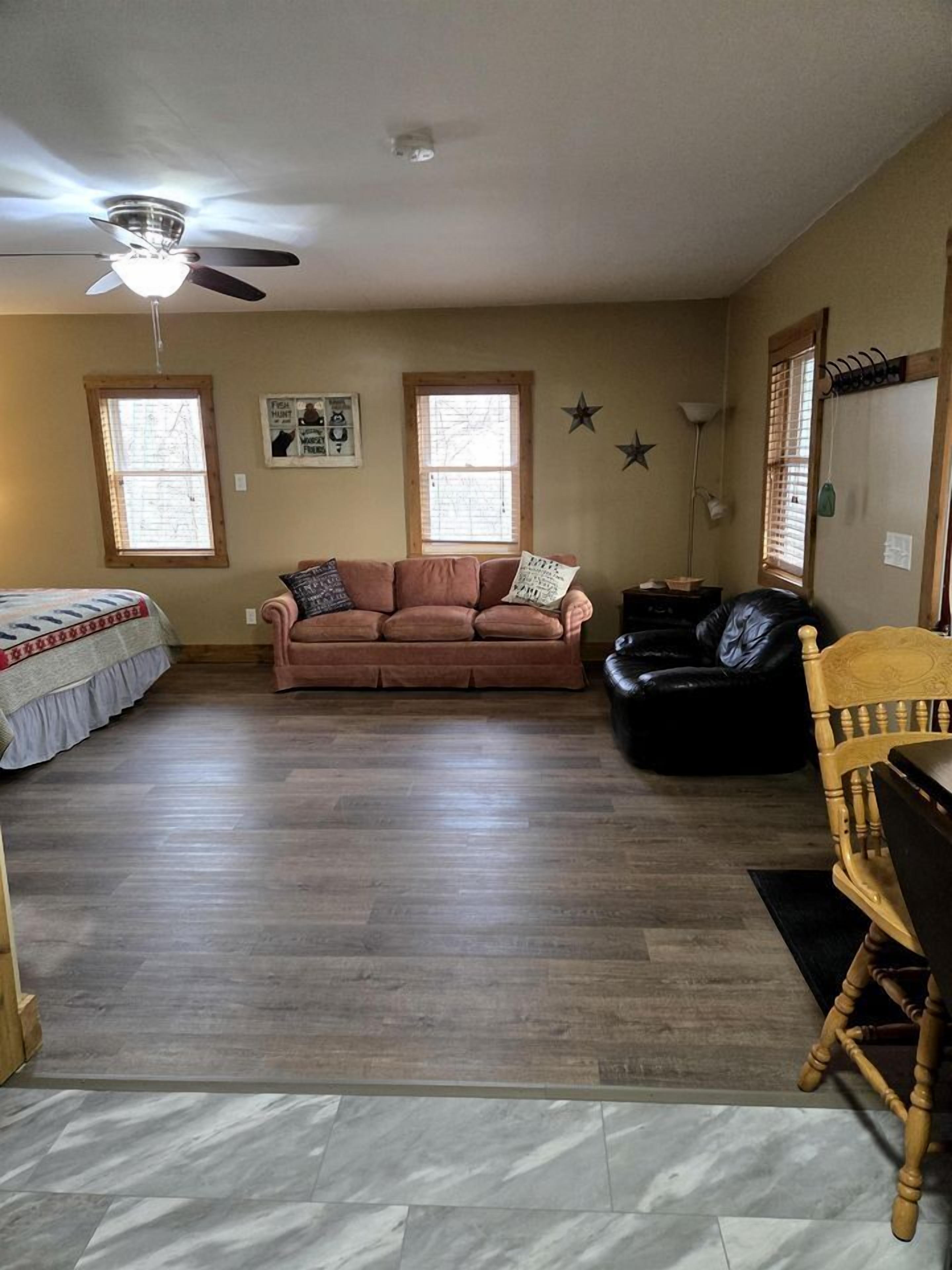 A living room with a ceiling fan, two windows, a sofa, a black leather chair, and a wooden chair.