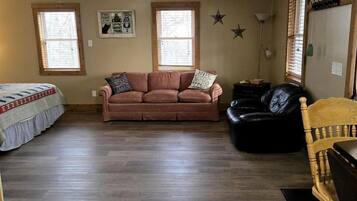 A living room with a ceiling fan, two windows, a sofa, a black leather chair, and a wooden chair.