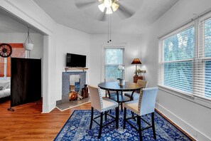 A dining area with a round table, chairs, a TV mounted on the wall, a fireplace, and a large window with blinds.